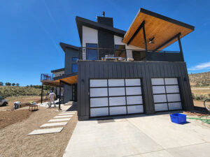 Modern two-story house near me with a sleek black and white exterior, boasting glass garage doors and a covered balcony. Under the clear blue sky, two people stand by the home.