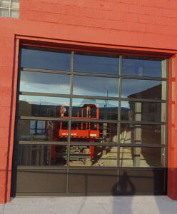A glass garage door with a sleek black frame graces the red brick building, reflecting a construction lift and the expansive sky above. It's a modern touch, right here near me.