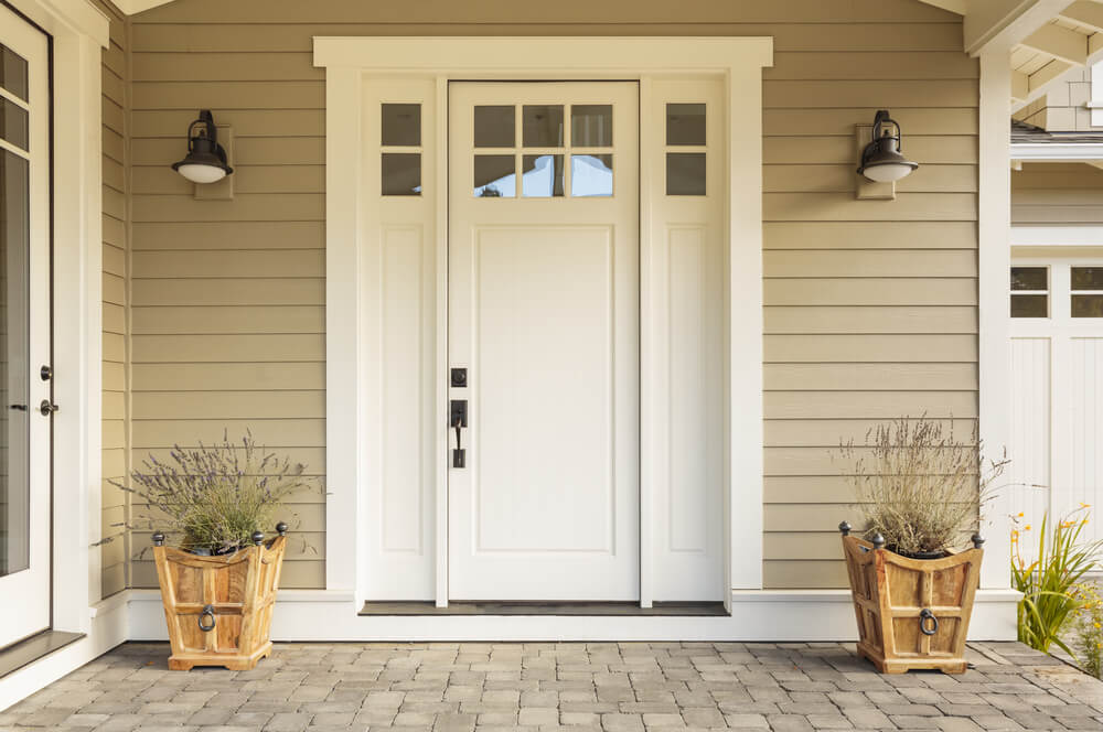White front door with glass panels, flanked by two potted plants in wooden containers—an inviting sight near me. Beige siding and wall-mounted lights accompany a charming brick pathway leading to the entrance.
