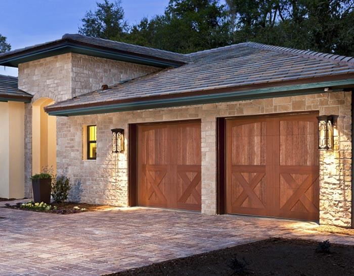A modern home exterior with stone walls, two wooden garage doors, outdoor lantern lights, and a paved driveway at dusk.
