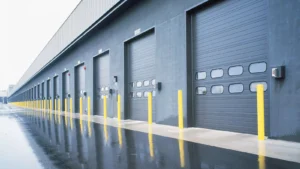 A row of closed loading dock doors with yellow safety bollards in front, next to a wet concrete driveway at an industrial warehouse.
