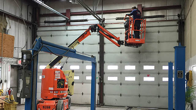 A worker operates an orange boom lift inside a garage, elevated to access the top section of a large roll-up door.