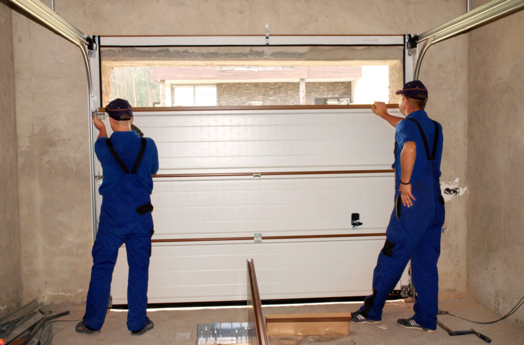 Two workers in blue overalls install a sectional garage door in an unfinished garage with bare walls.