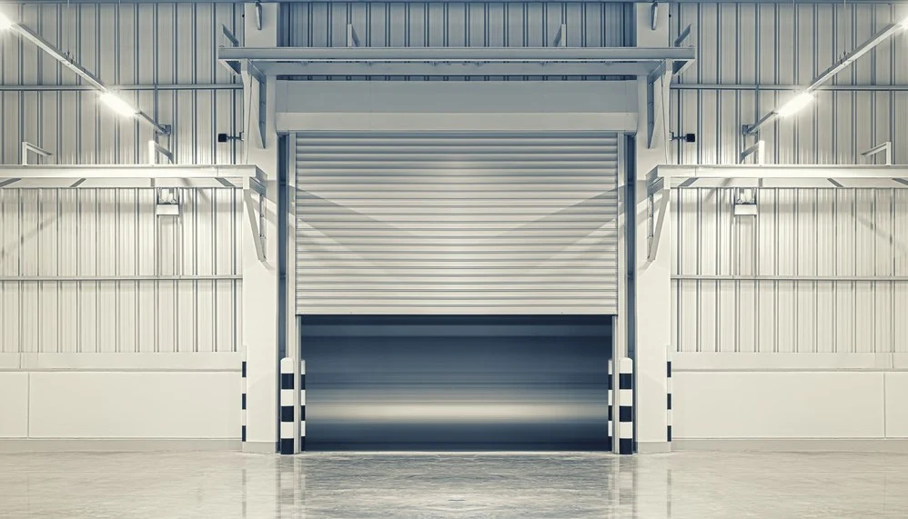 An empty industrial warehouse interior with white walls and a partially open large metal roll-up door, illuminated by overhead lights.