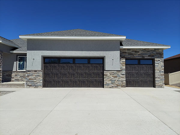 Modern house exterior with gray stucco, stone accents, and two dark wooden garage doors on a wide concrete driveway under a clear blue sky.