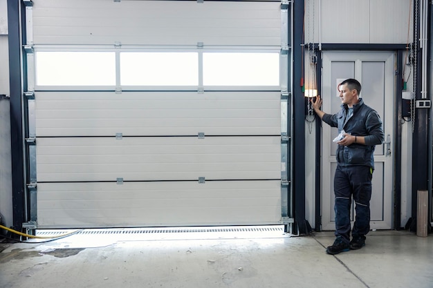 A man in work clothes stands near a wall, operating a control panel next to a large closed industrial garage door in a well-lit facility.