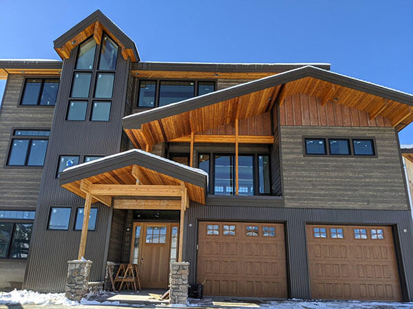 Modern three-story house with wood and metal exterior, large windows, two garage doors, and a covered entryway on a clear day.