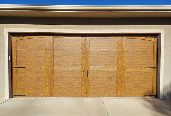 A closed wooden double garage door with two handles and visible hinges, set in a beige exterior wall under a light-colored roof trim.