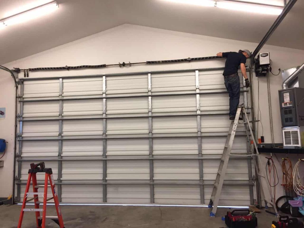 A person stands on a ladder inspecting or repairing the mechanism above a large closed garage door inside a well-lit garage.