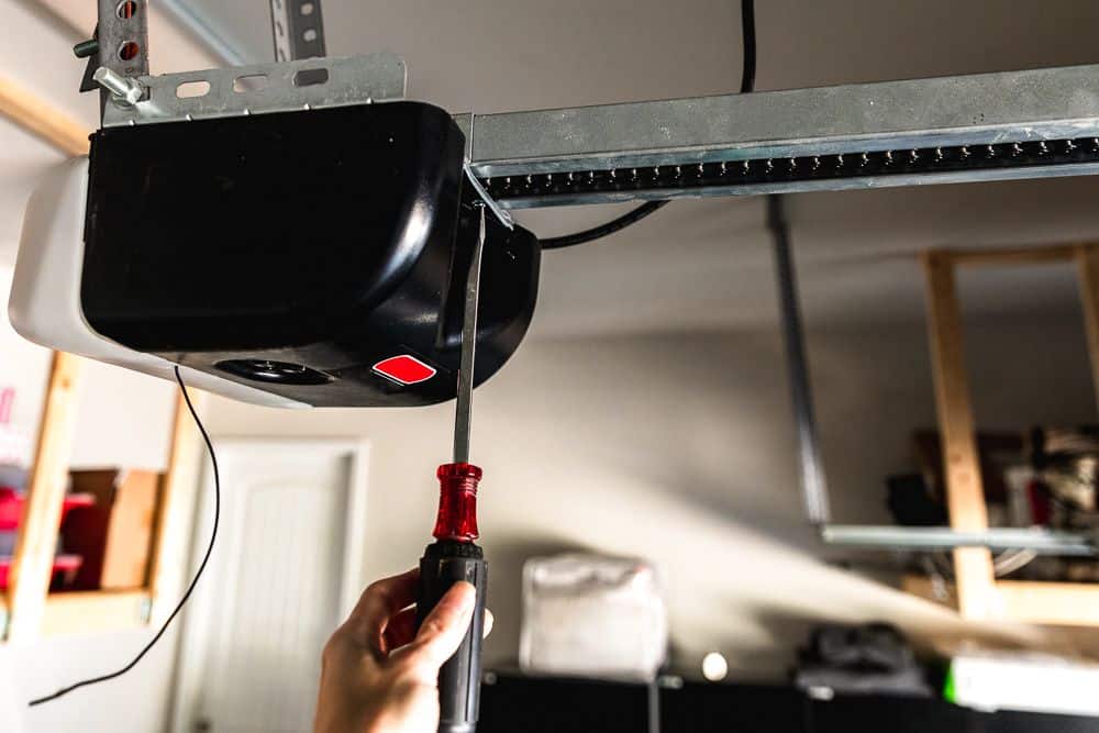 A person uses a screwdriver to adjust or repair a garage door opener mechanism mounted on the ceiling.
