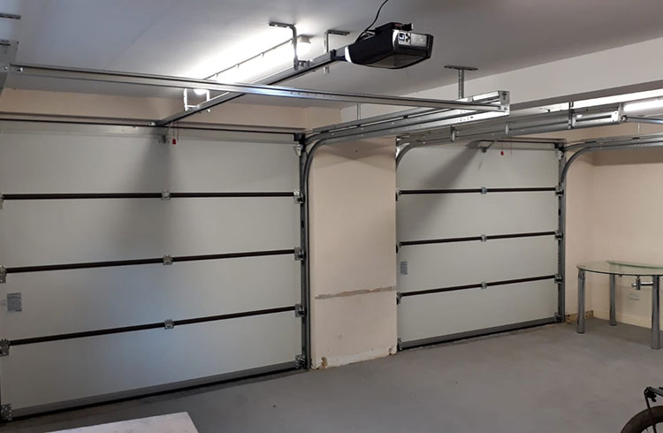 A clean, empty garage with two closed sectional garage doors, overhead tracks, fluorescent lighting, and a glass table in the corner.