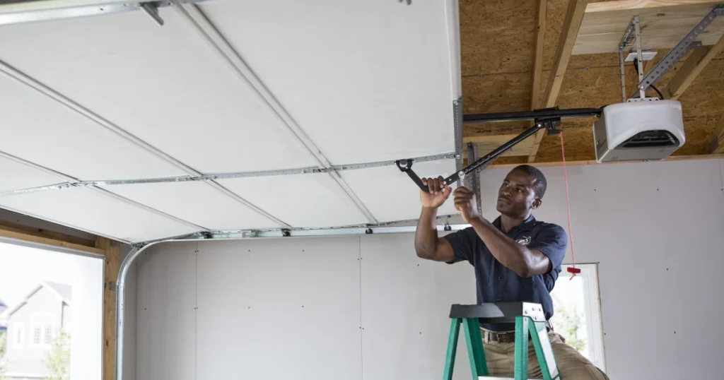 A man standing on a ladder repairs a garage door opener inside a garage.