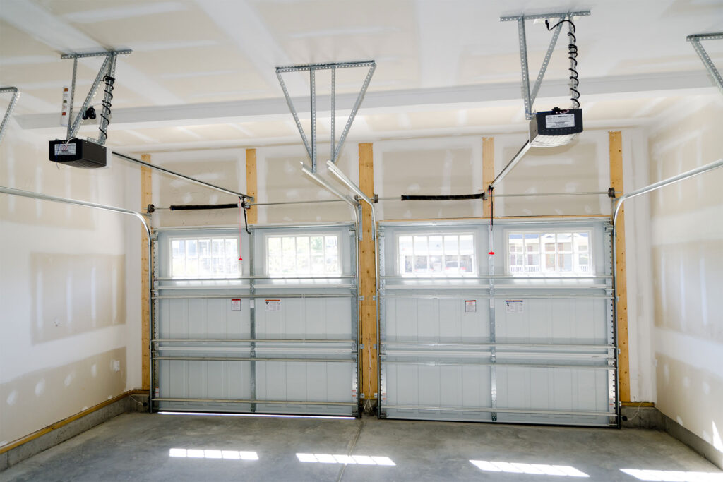 Interior view of a two-car garage with unfinished drywall, two closed garage doors with windows, and ceiling-mounted door openers.