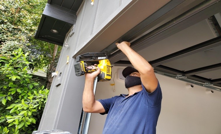 A person wearing a mask and blue shirt uses a power tool to work on the frame of a garage door.