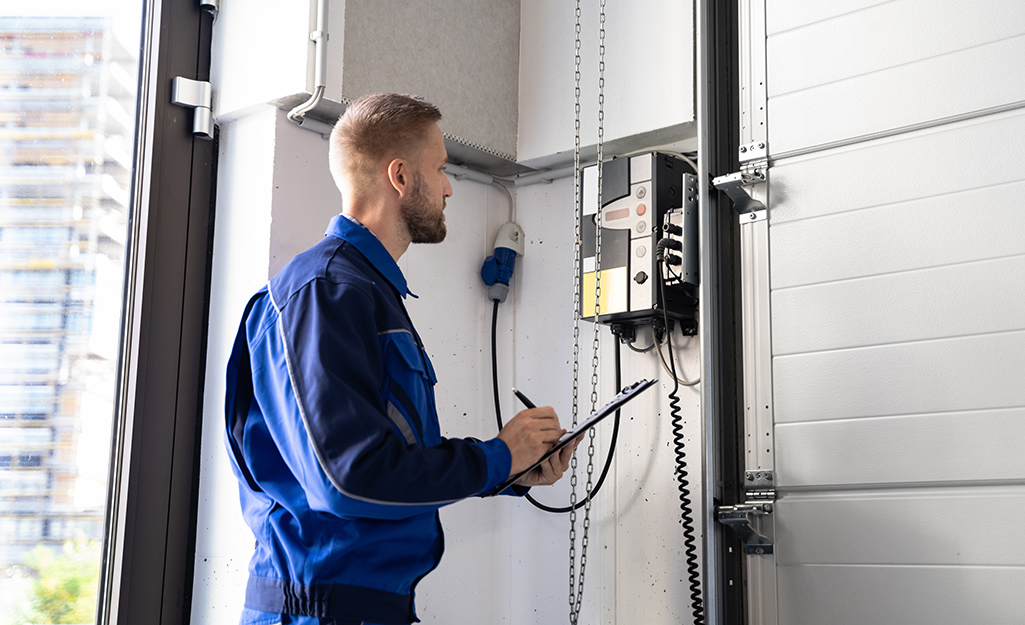 A technician in blue workwear inspects and records information from an electrical control panel mounted on a wall beside a large garage door.