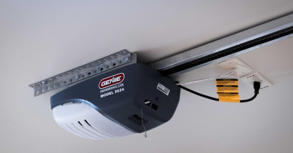 A Genie Model 3024 garage door opener is mounted on the ceiling with a metal rail and is plugged into a nearby electrical outlet.