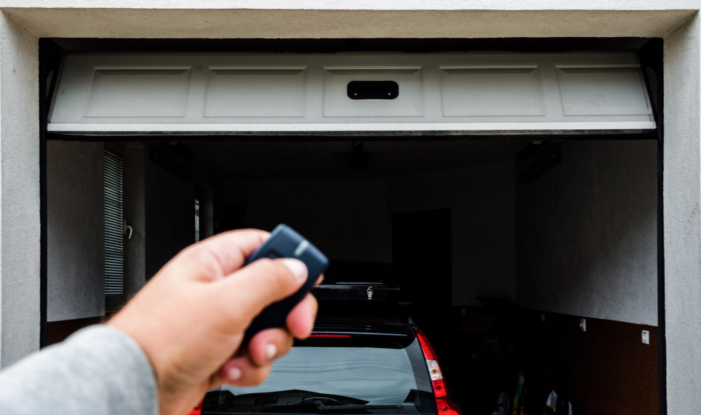 A person’s hand holding a remote control in front of a partially open garage door, with a car parked inside the garage.