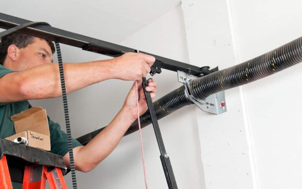 Person standing on a ladder installing or repairing a garage door opener mechanism with tools and components near the ceiling.