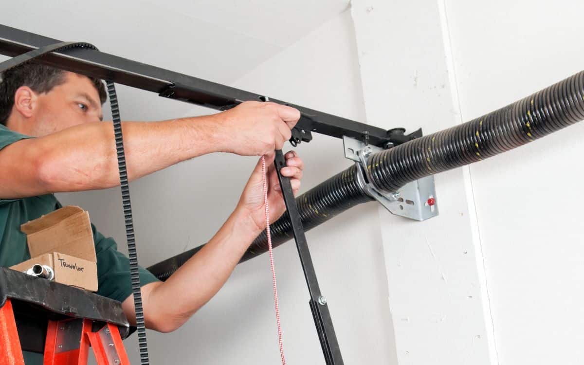 Person standing on a ladder installing or repairing a garage door opener mechanism with tools and components near the ceiling.