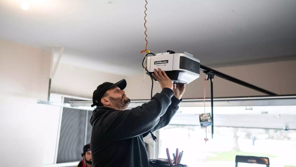 A man wearing a black cap and hoodie installs or repairs an automatic garage door opener, using tools while standing near the open garage.
