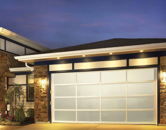 Modern house exterior at dusk featuring a large, frosted glass garage door with white grid framing, illuminated by wall-mounted lights.
