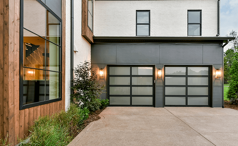 Modern house exterior with two frosted glass garage doors, black frames, wall-mounted lights, and large windows; concrete driveway and landscaped plants visible.