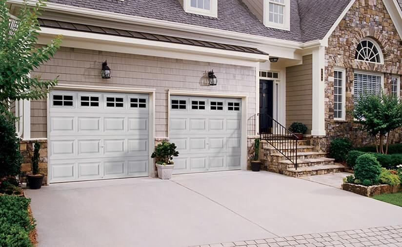 A suburban house with two white garage doors, stone and siding exterior, a front porch with steps, and landscaped shrubs along the driveway.