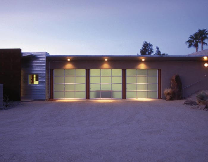 Modern single-story building at dusk with frosted glass garage-style doors, warm exterior lighting, a flat roof, desert landscaping, and palm trees in the background.